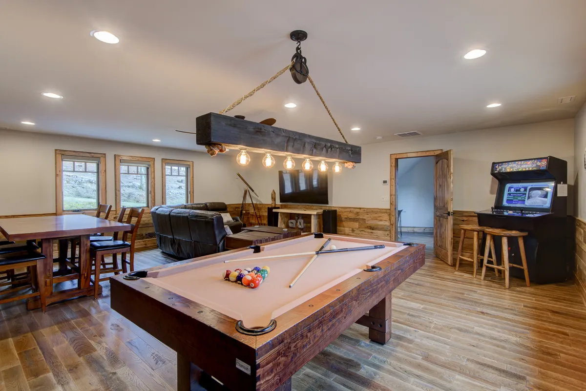 This inviting game room features a pool table at center stage, a retro arcade game machine, a charming modern chandelier, a spacious dining table, and plush leather seating. The layout encourages socializing, friendly competition, and relaxation, with a stylish yet playful vibe that sets the tone for fun indoor gatherings.
