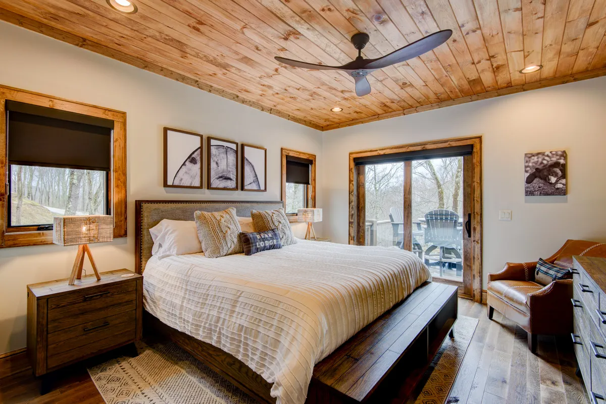 This image features a large bed with textured bedding situated under a wooden ceiling, bathed in natural light from expansive windows. A stylish armchair is positioned nearby, offering a serene corner for reading or quiet contemplation. The overall ambiance is tranquil, with soft light and a peaceful view outside.