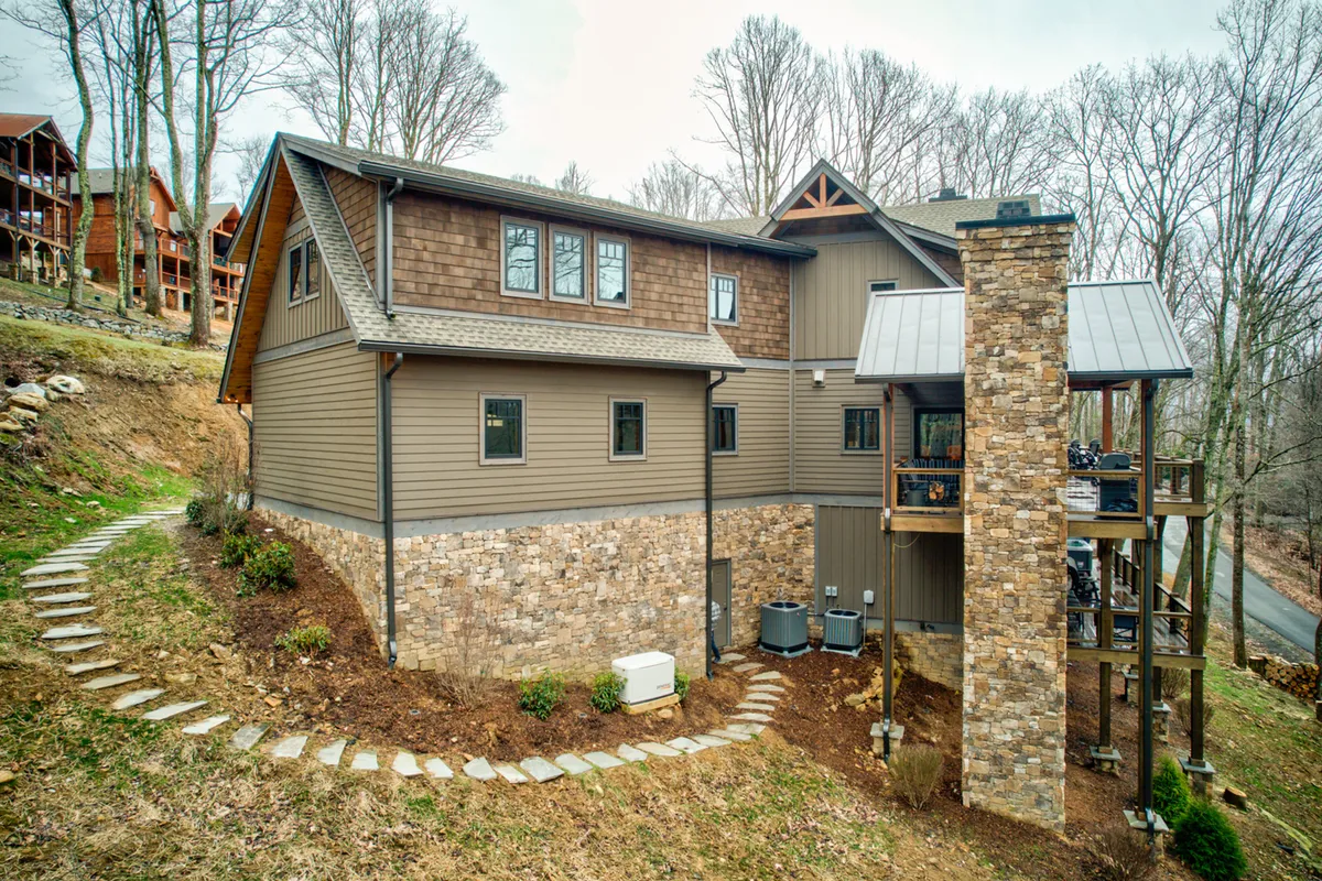 Featuring the wooden deck and stone chimney, this image draws attention to the outdoor gathering spaces, with a landscaped path inviting guests to explore. The scene suggests cozy evenings by the fire pit or lively family barbecues, set against nature's backdrop.