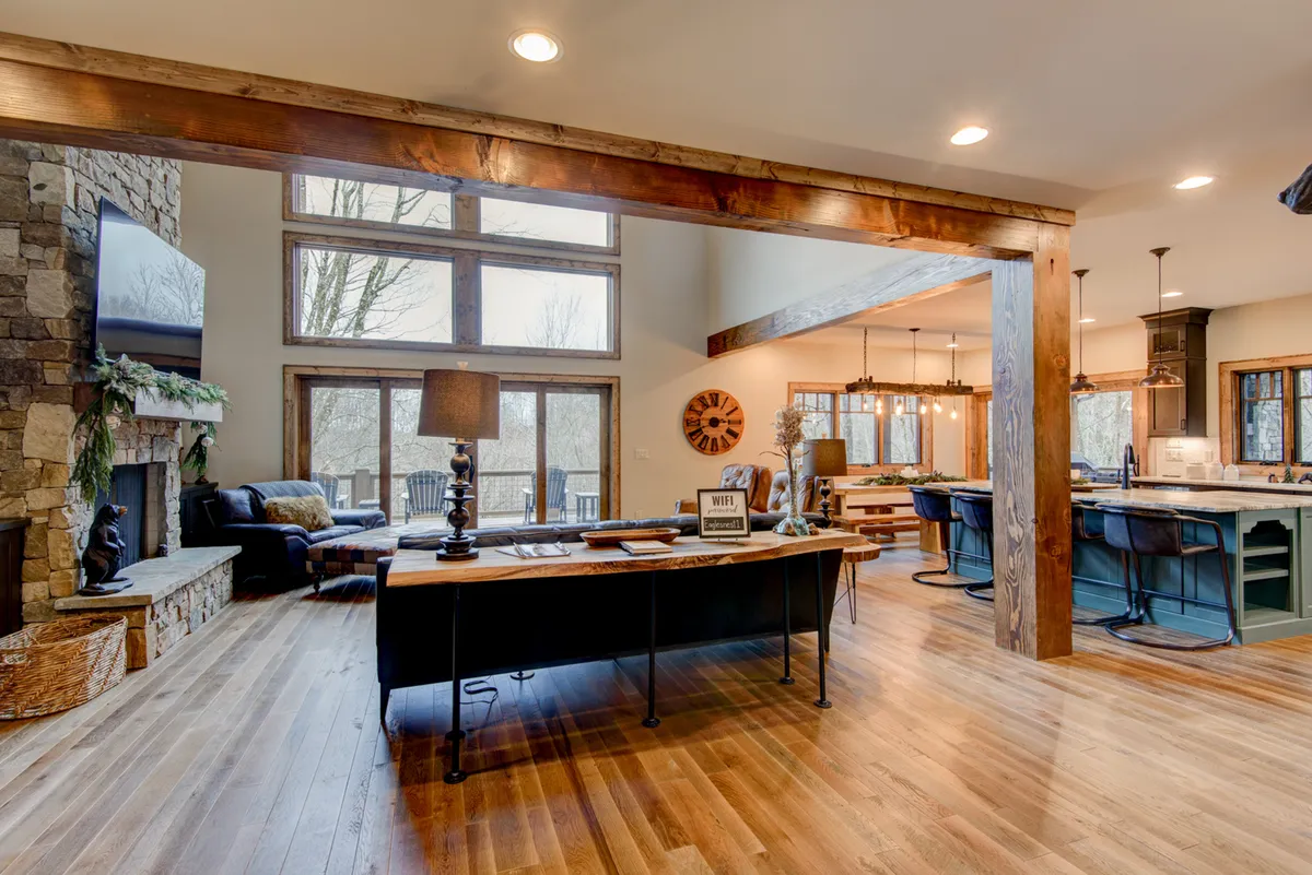 An inviting interior showcasing a large workspace area near the dining space, with a desk positioned close to large windows, stone fireplace, and wooden beams overhead. The workspace is functional yet stylish, ideal for productivity while remaining connected to the home’s warm environment.