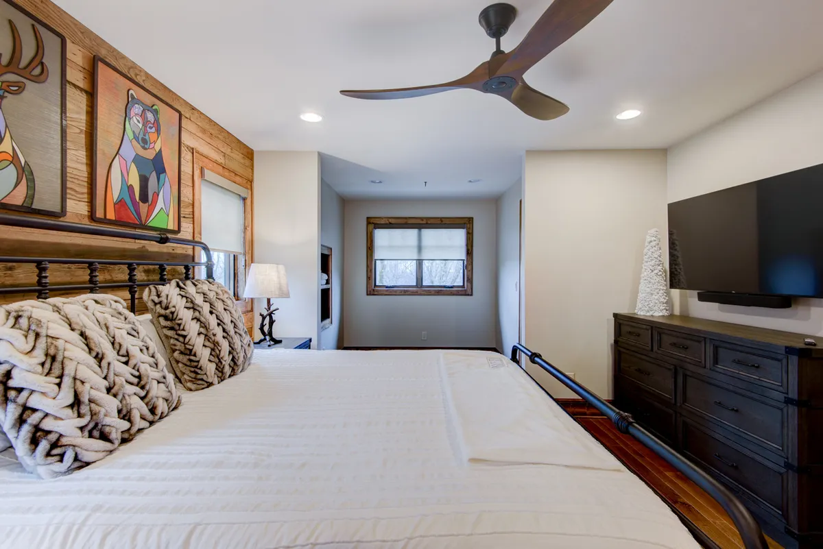 A cozy bedroom scene featuring a bed with plush pillows, tasteful artwork, and warm wooden furniture. The atmosphere is inviting and relaxing, with natural light filtering in and a television positioned for easy viewing from bed.