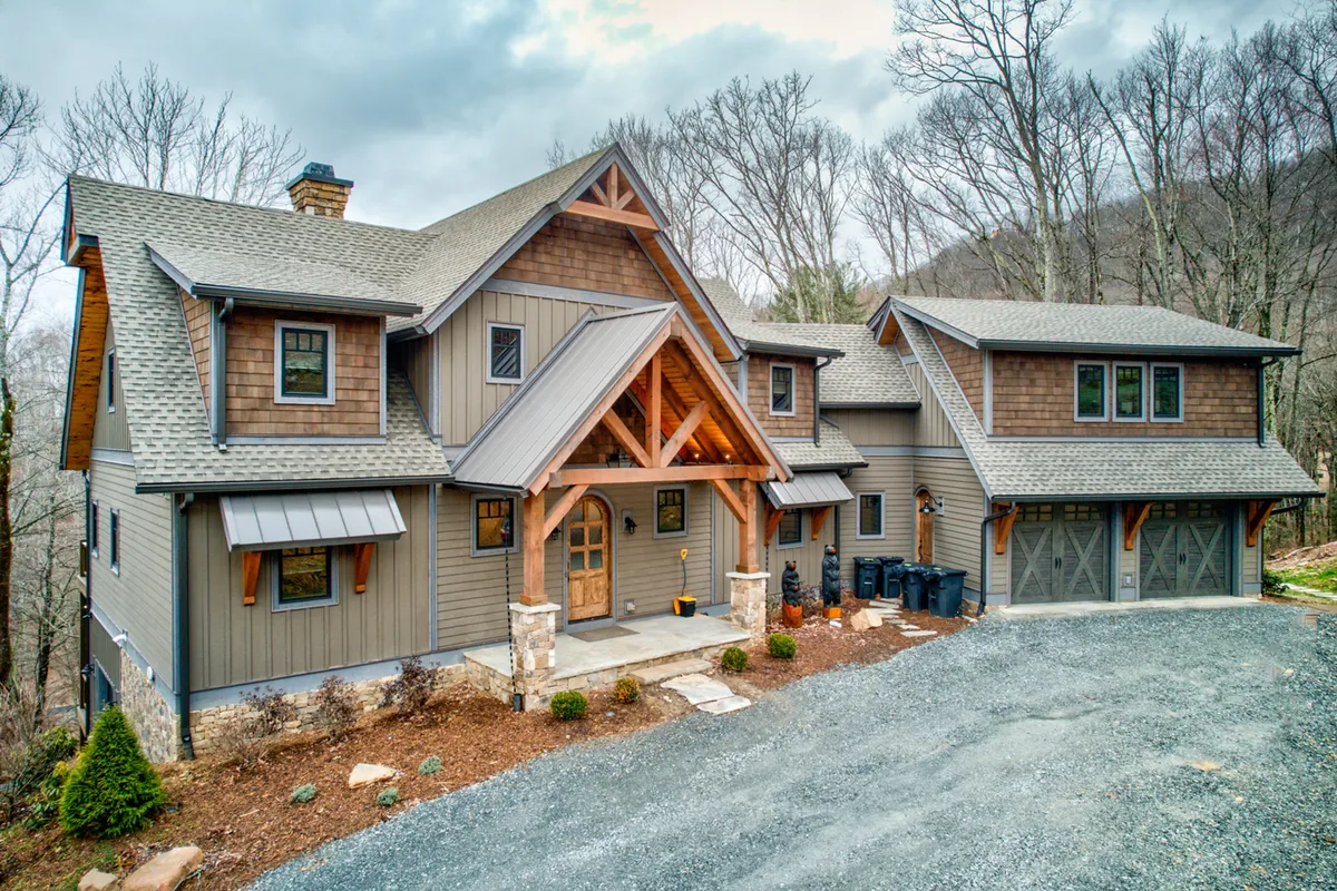 The image showcases the grand front entrance of Triple Bear Lodge, featuring a large wooden door framed by impressive stone pillars and an asymmetrical roofline. The house is surrounded by lush natural landscaping, evoking a sense of tranquility and seclusion. The inviting porch hints at cozy evenings spent watching sunsets or gathering with loved ones.