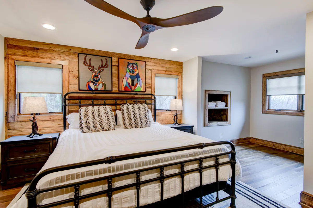 This image presents a calm and restful bedroom with a bed adorned with decorative pillows, framed artwork on the walls, and wooden nightstands. Large windows fill the space with natural light, creating a peaceful retreat ideal for relaxation or quiet moments with art.