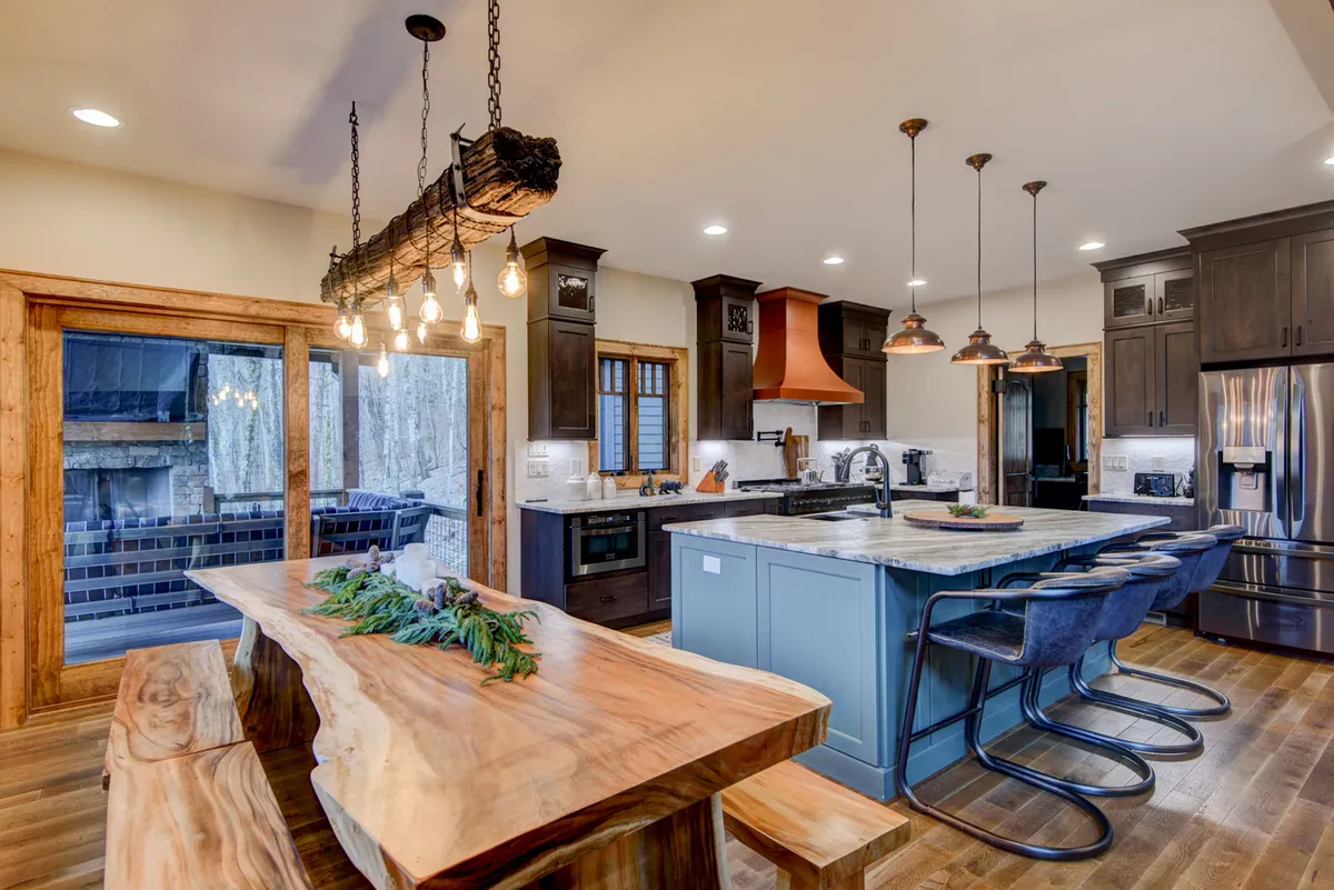 A beautifully set large wooden dining table sits adjacent to a stylish kitchen island with blue cabinetry. Overhead, pendant lights illuminate the area, and a striking copper hood complements the stove. The open layout and inviting décor make this the ideal spot for shared meals, celebrations, and culinary adventures.