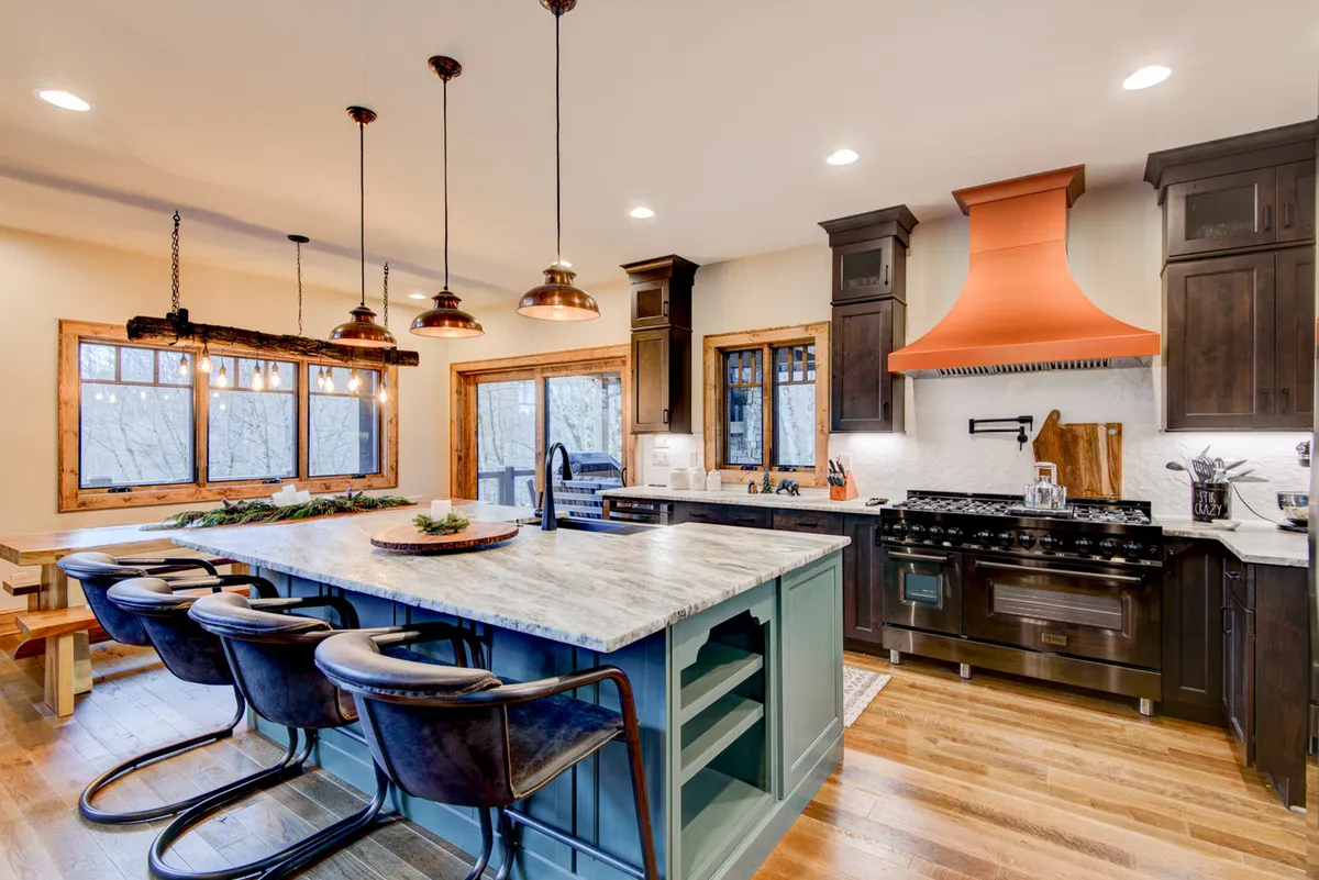 The kitchen is showcased with a large, marble-topped island, stylish dark cabinetry, a copper hood, hanging pendant lights, and expansive windows. The space is both functional and welcoming, encouraging group cooking, coffee mornings, and lively gatherings around the island.