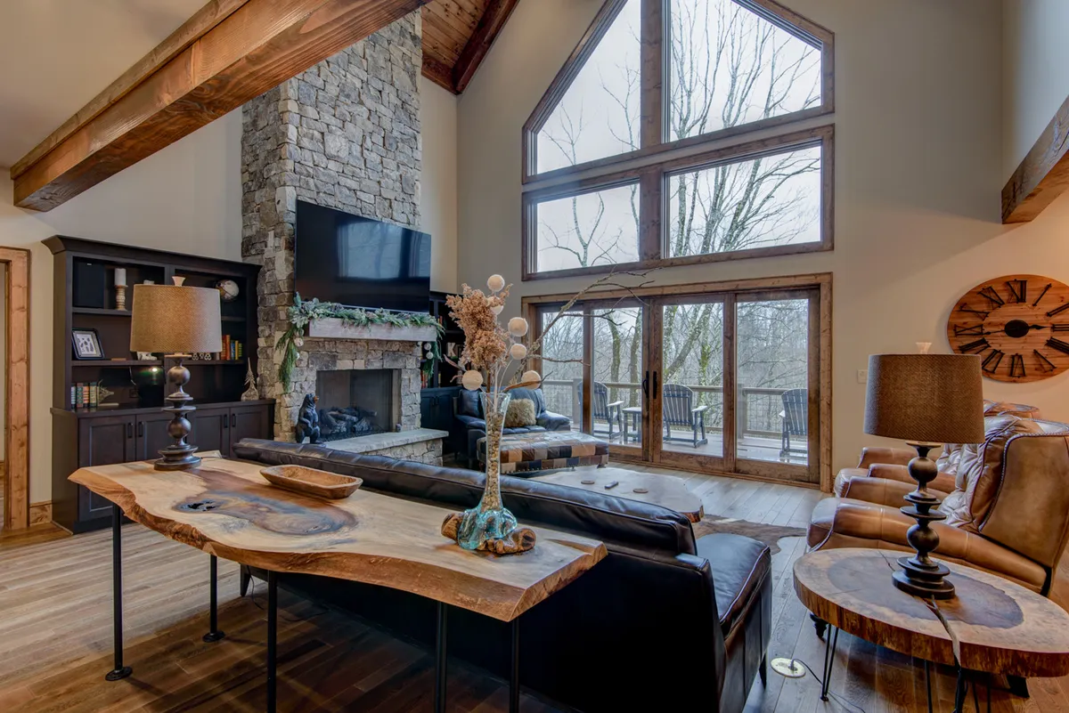 This image captures a spacious interior with a large stone fireplace as the focal point. Floor-to-ceiling windows flood the room with natural light, while leather sofas and a substantial wooden coffee table create a warm, inviting atmosphere. The space is clearly designed for relaxation, gathering, and conversation, with elegant finishes and a seamless flow between the living and dining areas.