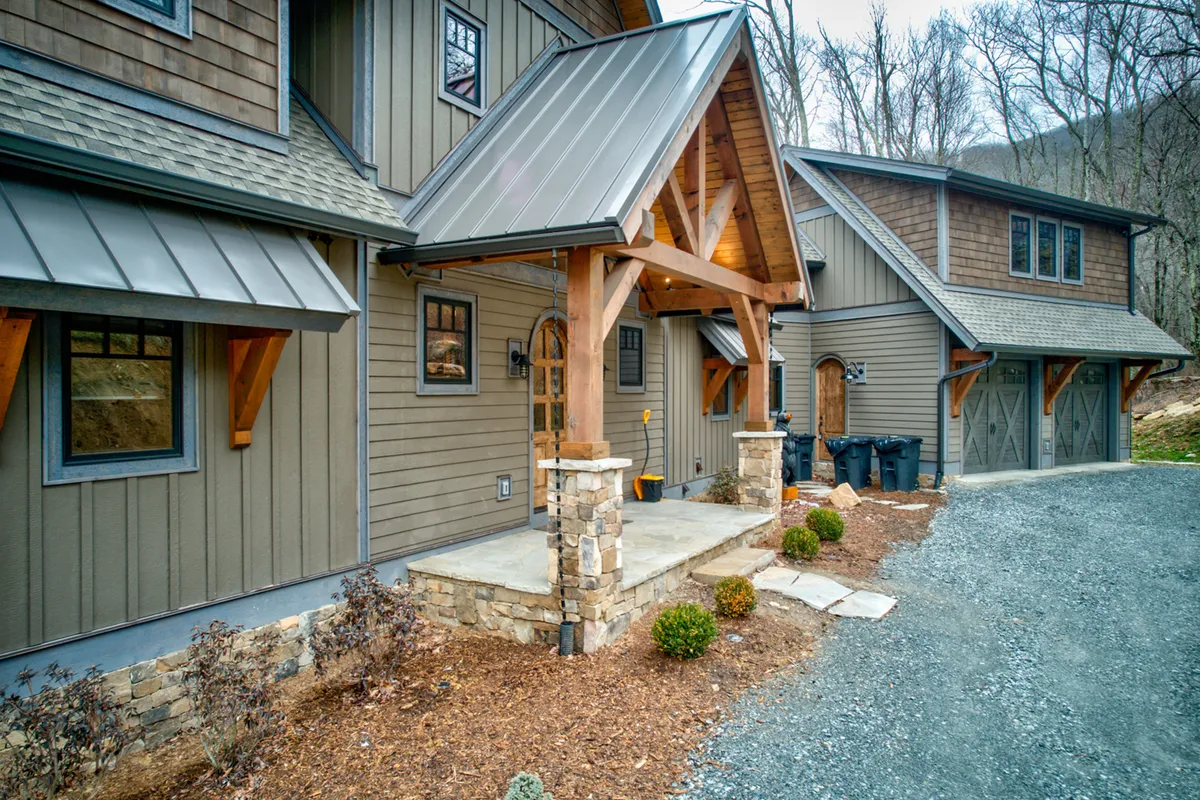 This image showcases the inviting front porch of Triple Bear Lodge, featuring sturdy wooden beams, stone pillars, a metal roof, and a beautifully landscaped garden area. The view hints at the expansive natural surroundings and the welcoming atmosphere guests experience upon arrival.
