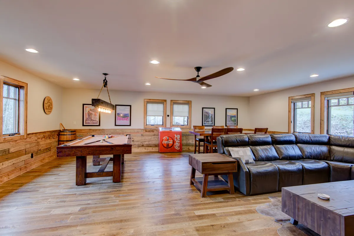 A spacious, casual entertainment area showcasing a pool table, a cozy leather sectional sofa, a dining table for board games or snacks, and a vintage Coca-Cola cooler. The room is designed for group activities, movie nights, and memorable gatherings, embodying the spirit of family fun and relaxation.