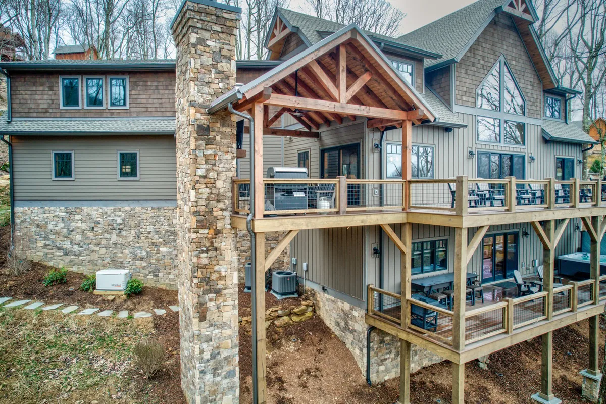 This image captures the exterior of Triple Bear Lodge, highlighting a stone chimney, wooden deck, large windows, and an inviting outdoor seating area. The setting exudes warmth, with hints of sunset views, making it ideal for outdoor dining, family gatherings, or relaxing while immersed in nature.