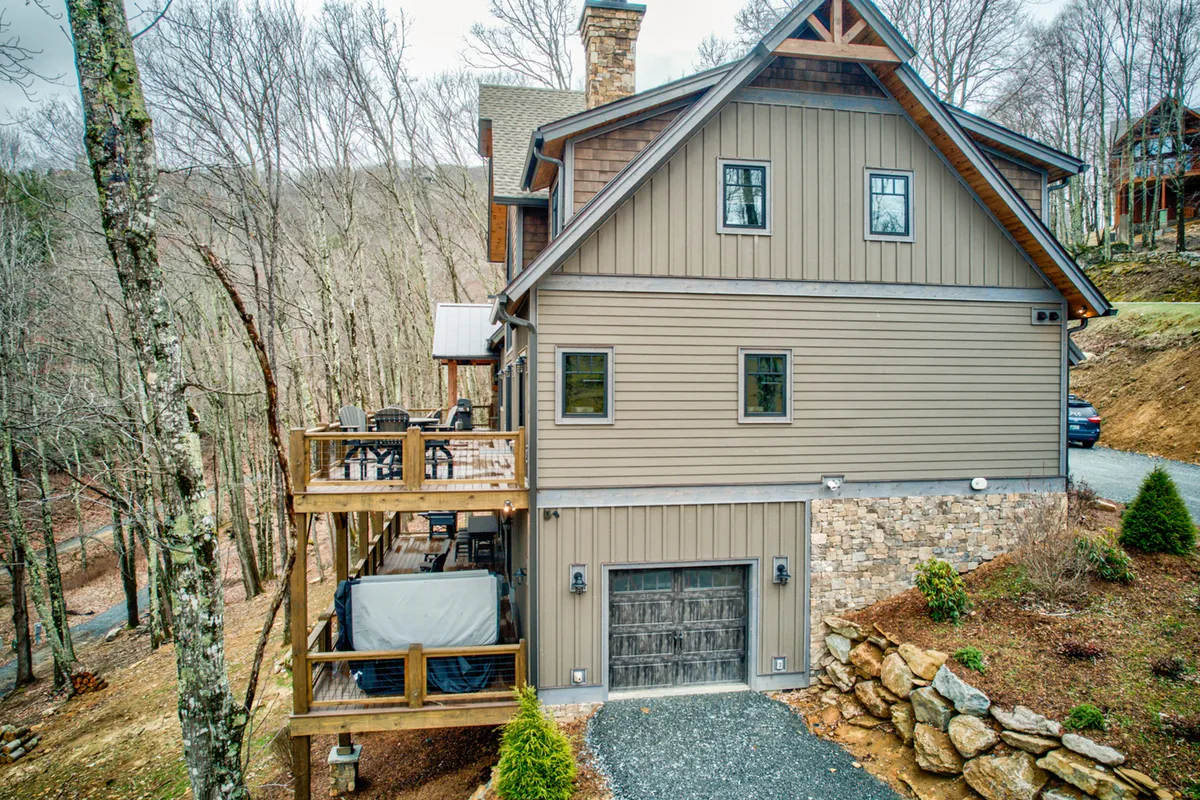 This exterior shot features a spacious wooden deck, stone garage, large windows, and is enveloped by trees. The scene is set for outdoor gatherings, relaxing on the deck, or sipping coffee in a peaceful, natural environment.