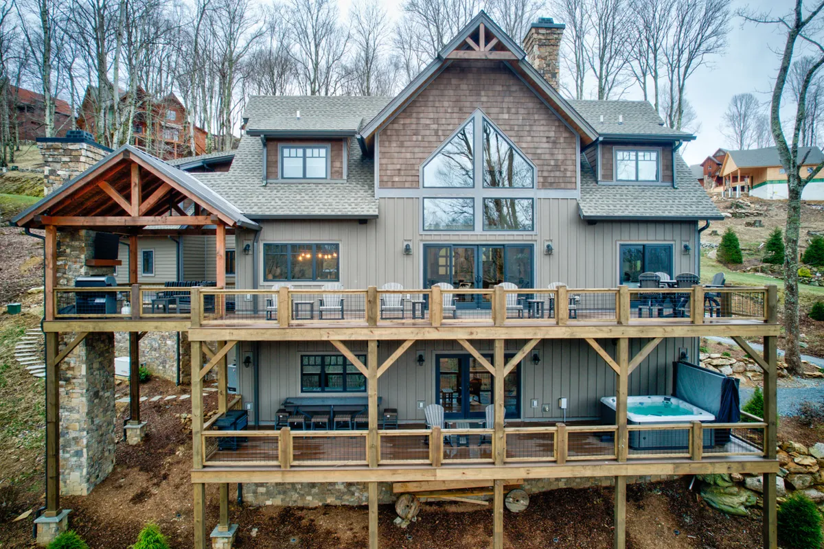 A welcoming exterior shot highlighting the large windows, an expansive wooden deck, a private hot tub, and elegant stonework, all set against a tranquil natural backdrop. The deck is furnished for outdoor dining and social gathering, creating a seamless flow between indoor and outdoor living spaces.