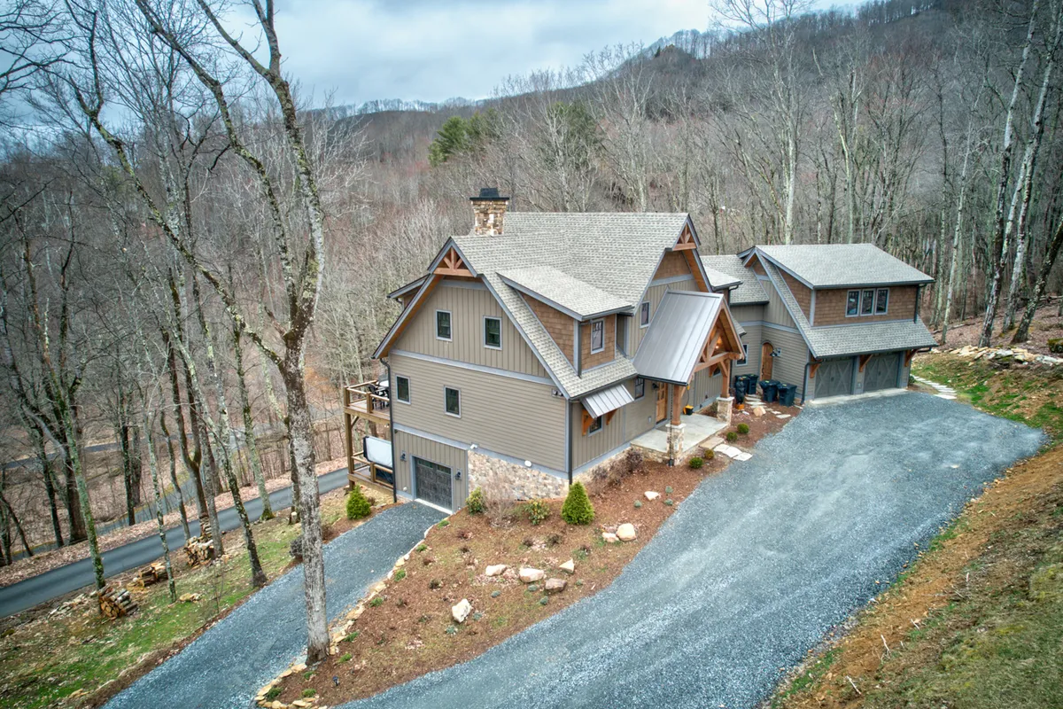 An exterior view of the home and its stone garage, set among leafy trees and a gravel driveway. The large outdoor area is ideal for backyard cornhole matches and social gatherings, and hints at the spaces where ping pong and firepit fun can unfold beneath the stars.